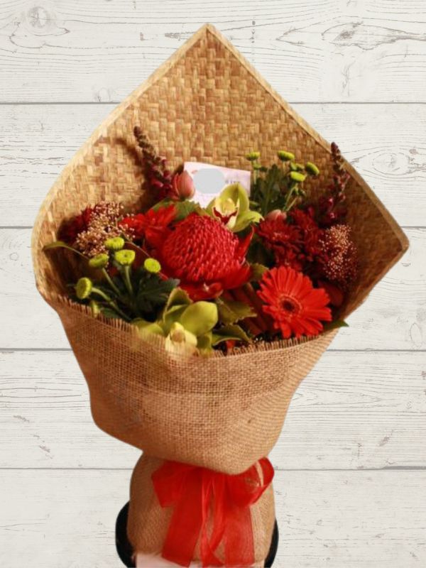 A fresh bouquet of red flowers wrapped in a harakeke mat, standing in a wooden vase on a wooden surface.