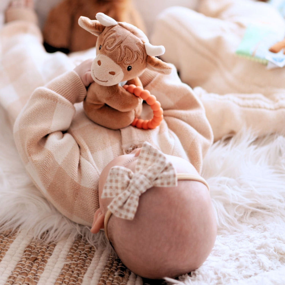 Baby wearing a checkered bow tie with a soft toy on a fluffy blanket with a highland cow rattle