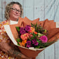 A person holding a hand-tied bouquet of hot pink and orange flowers, with seasonal variations in the flowers used.