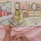 babys hands holding a paperback book with illustrations on it on a pink blanket