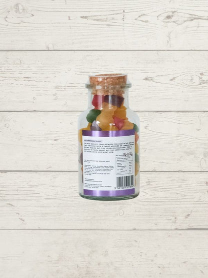 A clear glass jar filled with colorful gummy candies, sealed with a cork stopper and shown from the back with a white label, set against a light whitewashed wood plank background.