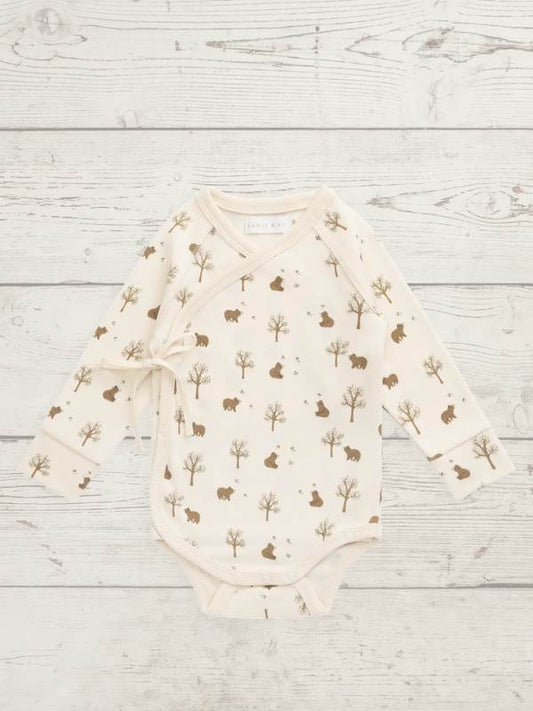 A long-sleeve baby bodysuit with an allover woodland print of brown trees, bears, and rabbits on a cream-colored fabric, laid flat on a light gray wood plank background.