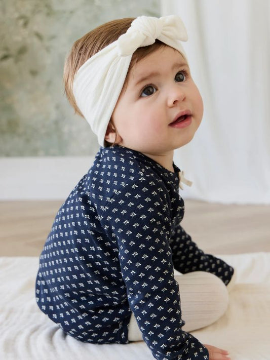 A baby wearing a navy long-sleeve top with a small white repeating pattern and a white knotted headband, sitting on a white blanket in a softly lit indoor setting.