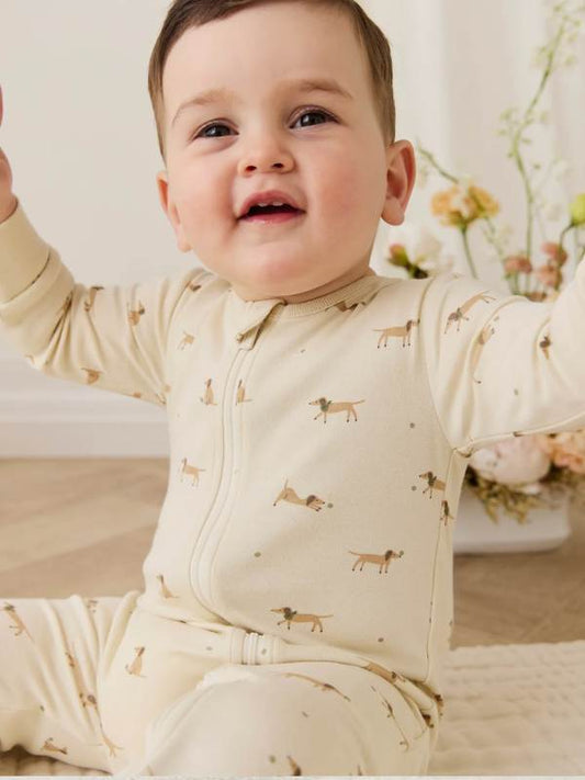 A baby sitting on a cream blanket , wearing a cream long-sleeve bodysuit with a small dog print.