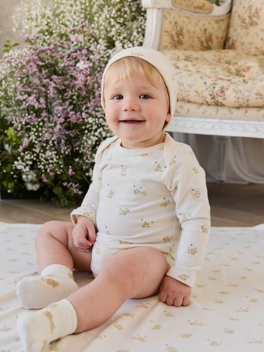 A baby sitting on a cream blanket in a decorated nursery, wearing a matching cream long-sleeve bodysuit with a small bunny print, a cream bonnet, and cream socks.