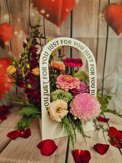 A colorful floral arrangement with a variety of flowers and greenery, displayed in a container with the text 'JUST FOR YOU' on it, against a wooden backdrop with heart decorations.