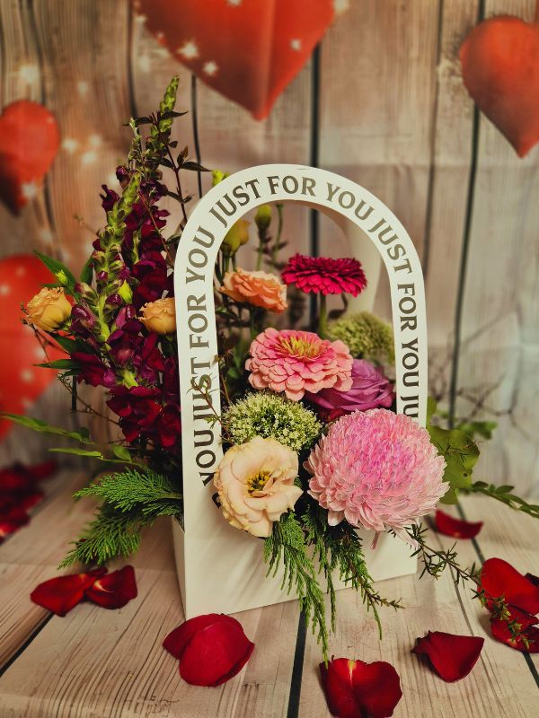 A colorful floral arrangement with a variety of flowers and greenery, displayed in a container with the text 'JUST FOR YOU' on it, against a wooden backdrop with heart decorations.