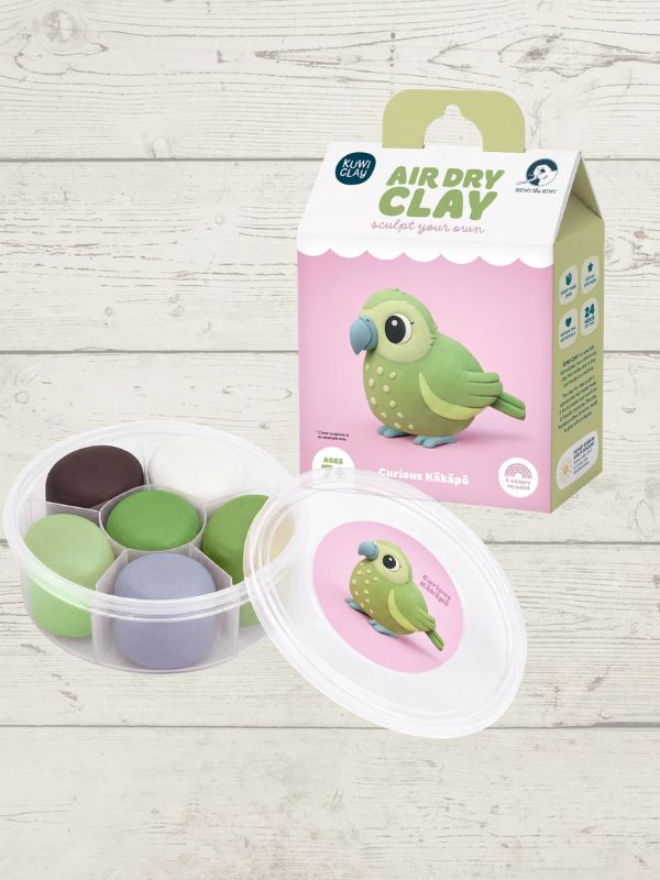 A children's air dry clay kit shown with its retail box and a clear plastic container of multiple colored clay pieces on a light wood plank background. The box is pink and green with a cartoon parrot, and the container holds several small clay balls in different colors.