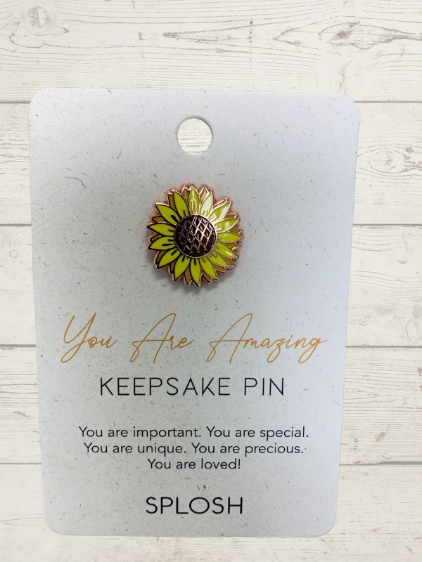 A keepsake pin with a sunflower design, accompanied by a message card stating 'You are Amazing' and 'You are loved'. The pin is displayed against a wooden background.