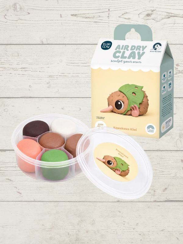 A children's air dry clay kit shown with its packaging box and a clear plastic container holding multiple colored clay pieces, placed on a light wood plank background.
