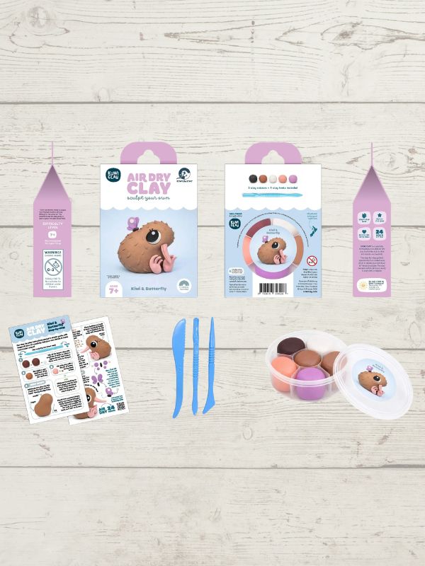 A clay sculpting kit laid out on a light wood background, including two pink-and-white hanging boxes, two instruction sheets, three blue sculpting knives, and a small round container with colorful clay pieces and a clear lid.