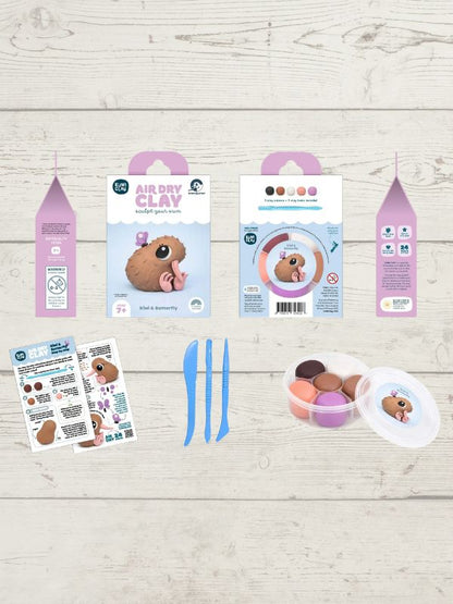 A clay sculpting kit laid out on a light wood background, including two pink-and-white hanging boxes, two instruction sheets, three blue sculpting knives, and a small round container with colorful clay pieces and a clear lid.