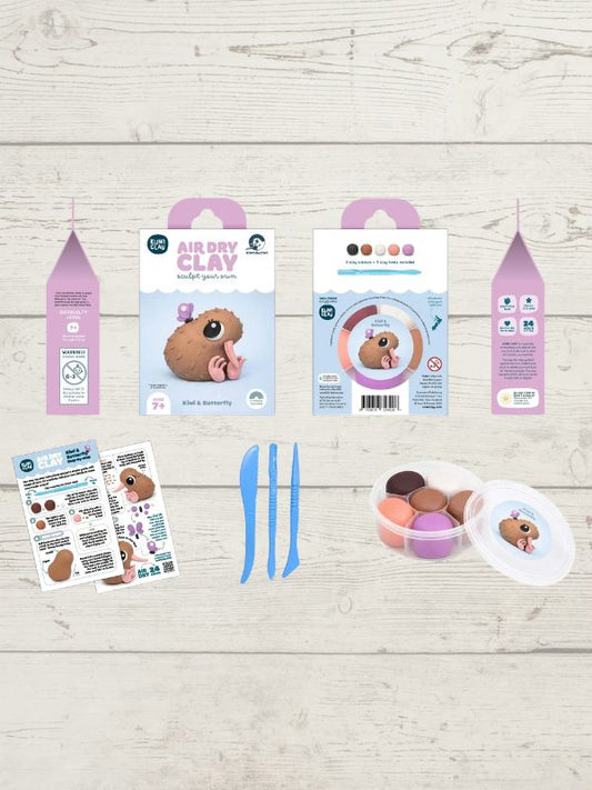 A clay sculpting kit laid out on a light wood background, including two pink-and-white hanging boxes, two instruction sheets, three blue sculpting knives, and a small round container with colorful clay pieces and a clear lid.