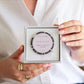 Person holding a box with a bracelet labeled 'Amethyst' against a white background