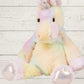 A multicolored plush toy resembling a unicorn with a white body, pink ears and tail, and yellow and green accents on a wooden background.