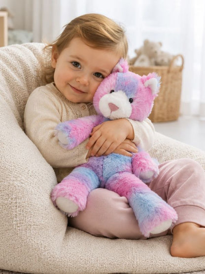 A young child sitting in a cream-colored plush beanbag chair, holding a pastel rainbow plush bear toy. The child is wearing a cream knit sweater and pink pants, with a soft, bright indoor room in the background.