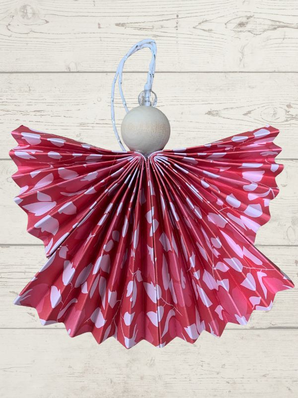 A festive angel Christmas ornament featuring a red and white patterned pleated paper skirt, a light wood ball torso, and a light blue hanging loop, set against a light wood plank background.