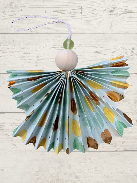 A handmade paper angel ornament with a light blue background, decorated with gold, brown, and green watercolor-style spots. It features a pleated paper body, a light wood bead, a green bead, and a white hanging string against a light wood plank background.