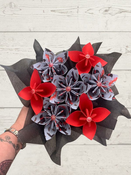 A bouquet of artificial flowers made from folded gray patterned paper and bright red paper petals, wrapped in dark gray tissue paper and held by a tattooed hand against a white wood plank background.