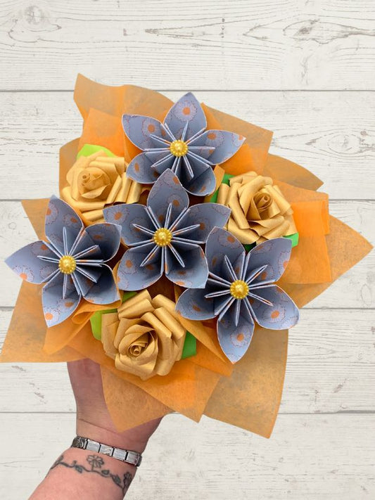 A hand holding a bouquet of origami paper flowers, featuring blue-gray paper flowers with orange spots and yellow centers, alongside tan paper roses, wrapped in orange tissue paper against a white wood background.