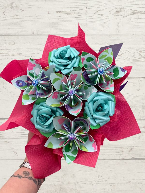 hand holding a paper posy box of flowers in teals, pinks and greens