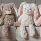 Two soft toy bunnies with beige and white coloring, sitting on a wooden surface. One bunny is plush and the other is a stuffed toy, both with embroidered eyes and a textured surface.