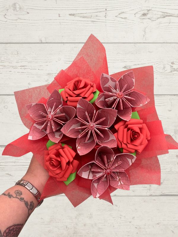 A hand holding a bouquet of red and pink origami paper flowers, including roses and starburst blossoms, wrapped in red tissue paper against a white wood plank background.