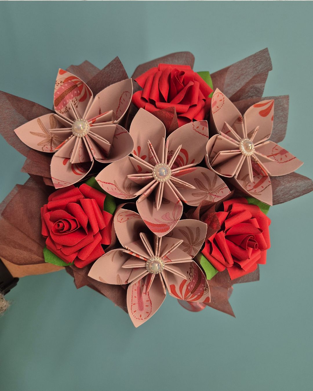 A bouquet of handmade origami flowers, featuring red paper roses and beige paper blossoms with patterned details, wrapped in brown tissue paper against a solid teal background.