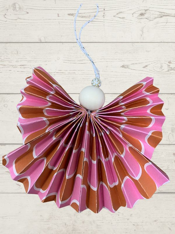 A handmade paper angel ornament with pink, orange, and white patterned wings, a white bead body, and a blue hanging string, shown against a light wood plank background.