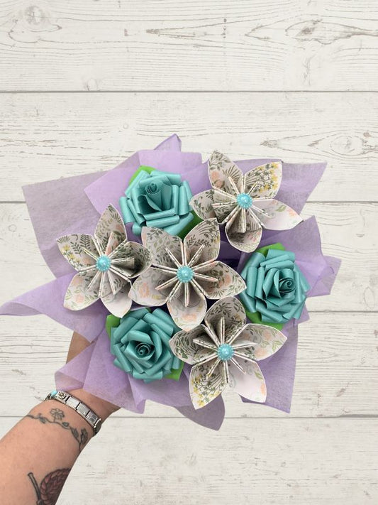 A hand holding a bouquet of paper flowers made from book pages, featuring teal paper roses and patterned paper flowers wrapped in light purple tissue paper, set against a white wood plank background.
