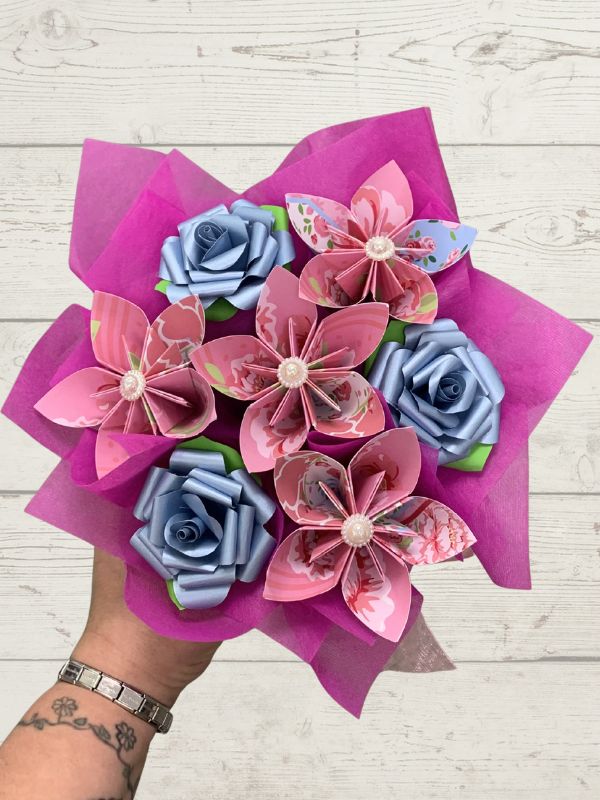 Bouquet of pink and blue paper flowers held by a person on a light wooden surface.