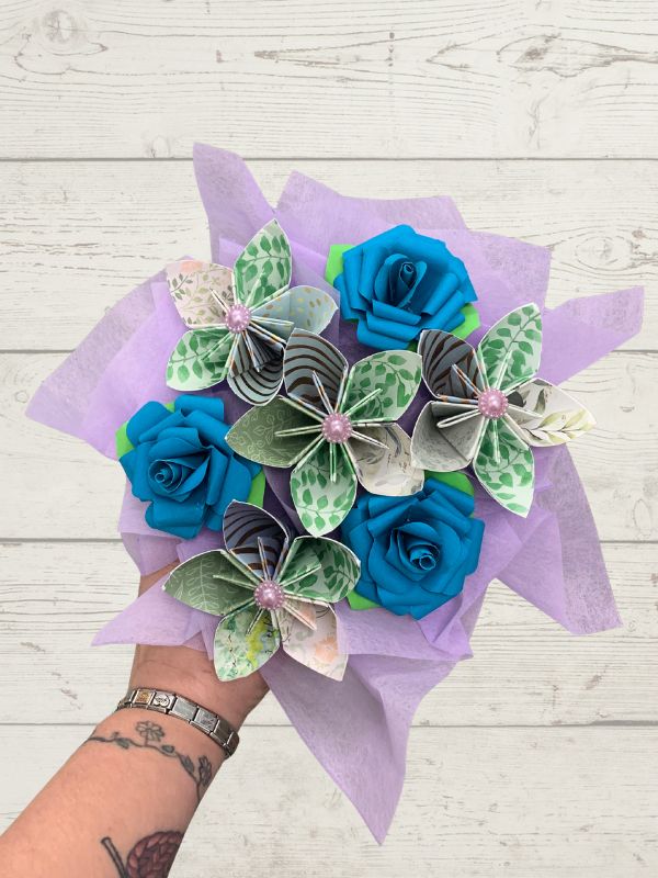 A hand holding a bouquet of artificial flowers made from blue satin roses and green-and-white patterned paper blossoms, wrapped in light purple tissue paper against a white wood plank background.