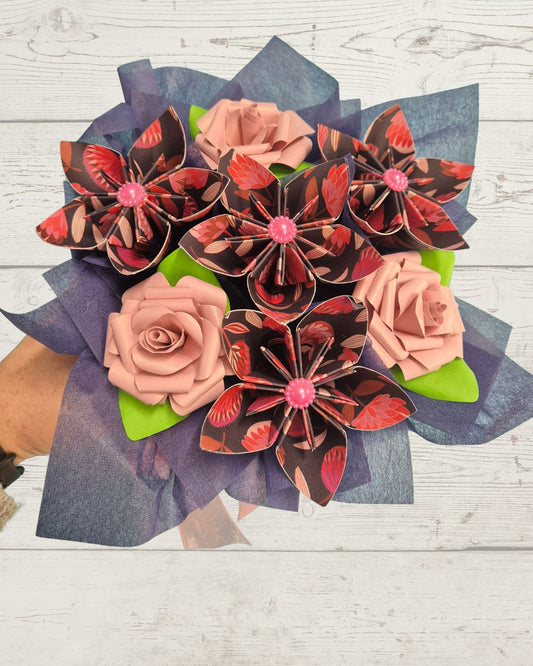A handmade bouquet of origami flowers, featuring patterned red and black paper blossoms with pink centers and peach paper roses, wrapped in blue-gray tissue paper and held by a hand against a white wood background.