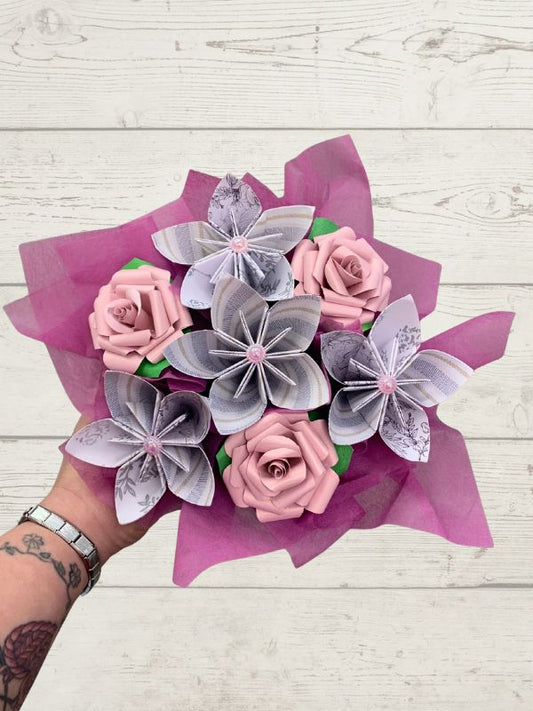 A hand holding a bouquet of artificial flowers made from pink paper roses and folded paper flowers, wrapped in magenta tissue paper, against a white wood plank background.
