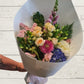 A bouquet of fresh florals in apricots, pinks, blues and whites wrapped in white paper with a white box.