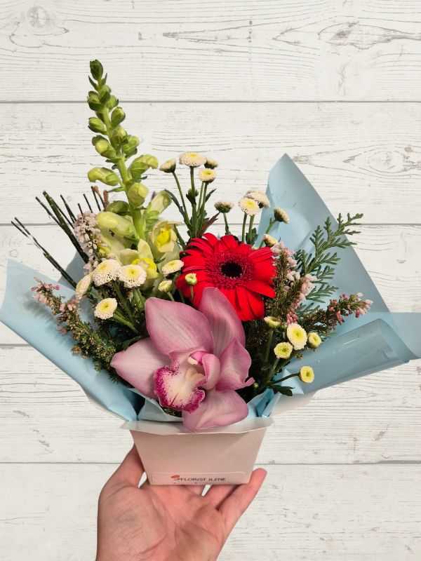 Bouquet of flowers with pink orchids and red gerbera daisies held against a light wooden background