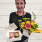 person holding a floral arrangement, cupcakes and a handmade card. 