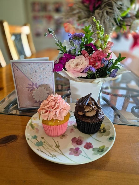 A fresh flower posy with two cupcakes and a handmade card displayed on a table.