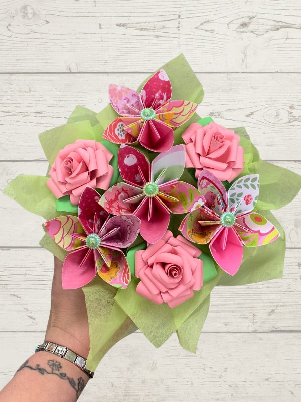 A hand holding a bouquet of colorful origami paper flowers, including pink roses and butterfly designs, wrapped in light green tissue paper against a white wood plank background.