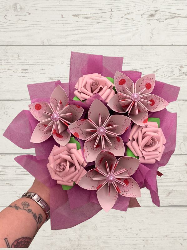 A bouquet of handmade pink origami flowers, including roses and lilies, wrapped in magenta tissue paper and held by a tattooed hand against a white wood background.