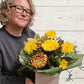 Person holding a posy box of yellow flowers against a light wooden background