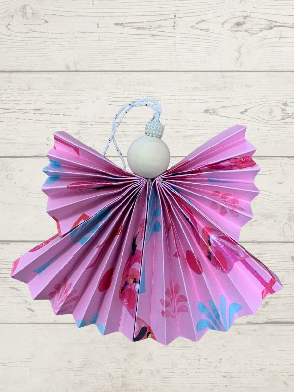 A pink paper angel ornament with a pleated skirt, floral pattern, and a white wooden head hanging from a blue string, set against a light wood plank background.