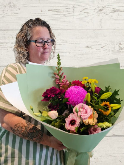 A person holding a bouquet of flowers including snapdragons, sunflowers, chrysanthemums, lisianthus, wrapped in white and green paper. 