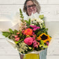 A bouquet of assorted fresh flowers including pink roses, sunflowers, and other seasonal flowers, arranged in a white paper vase with a yellow bow.