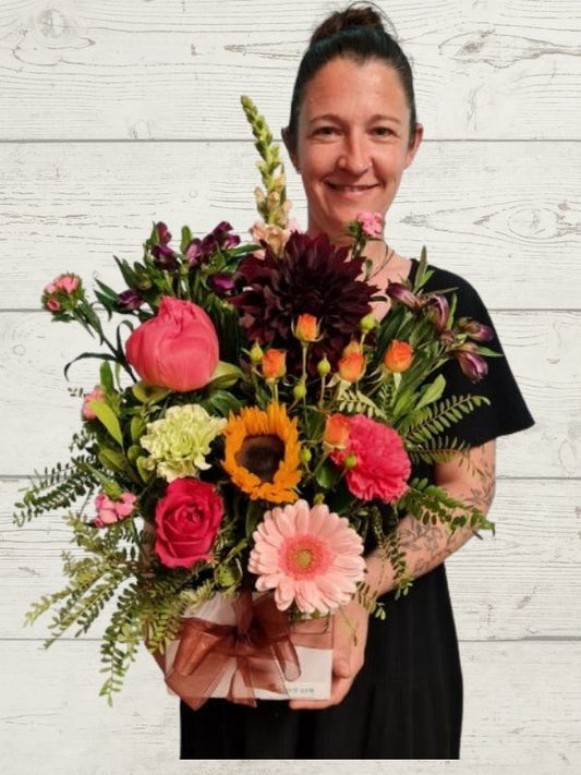 A person holding an arrangement of flowers including peonies, sunflowers, gerberas, roses, dahlias arranged into a white box with a golden ribbon bow. 