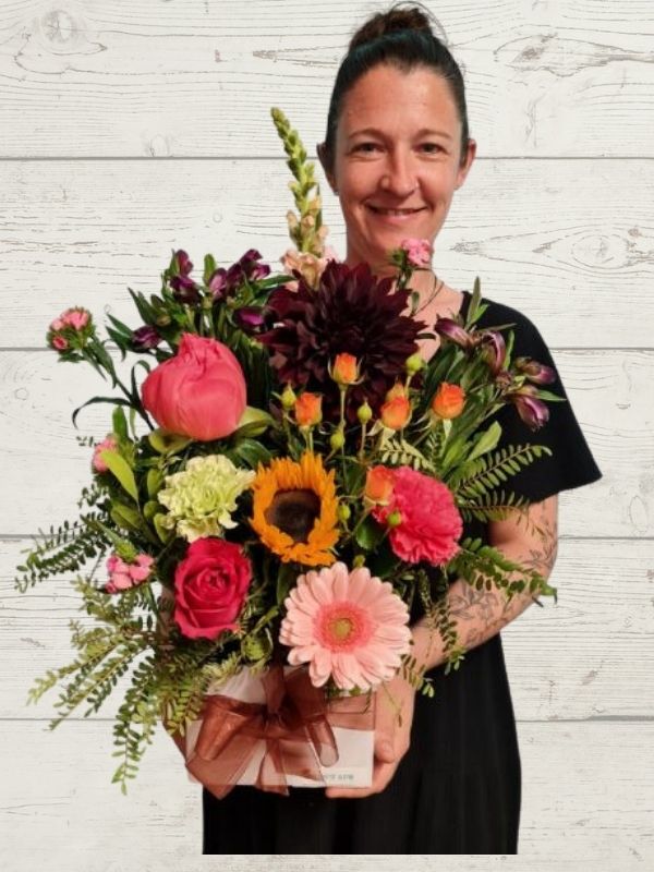 A person holding an arrangement of flowers including peonies, sunflowers, gerberas, roses, dahlias arranged into a white box with a golden ribbon bow. 