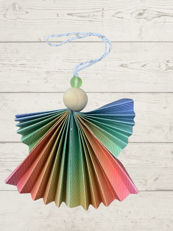 A hanging decorative angel ornament made from folded paper in a rainbow gradient, with a light wood bead torso, a green bead head, and a blue hanging string, shown against a whitewashed wood plank background.