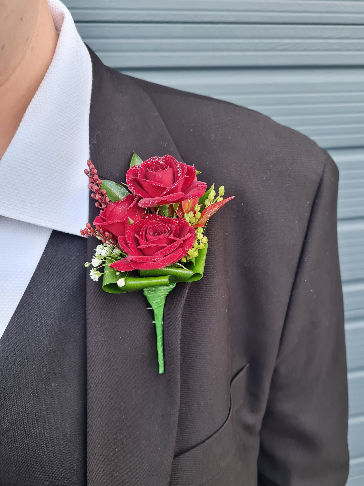 A close-up of a men's formal wear featuring a dark brown suit jacket, white dress shirt, and a vibrant red rose boutonniere with green leaves and small white filler flowers.