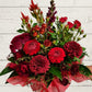 A red-themed floral arrangement featuring various red flowers and green foliage, arranged in a container with red wrap.