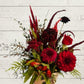 A red, brown, and green floral arrangement with various flowers including shades of red and a few green leaves, held against a white wooden background.